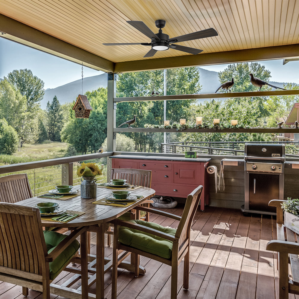 Outdoor smart ceiling fan with light installed on a covered patio, featuring walnut and Brazilian cherry dual-finish blades, above a cozy seating area with stone flooring and garden views. 