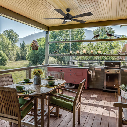 Outdoor smart ceiling fan with light installed on a covered patio, featuring walnut and Brazilian cherry dual-finish blades, above a cozy seating area with stone flooring and garden views. 