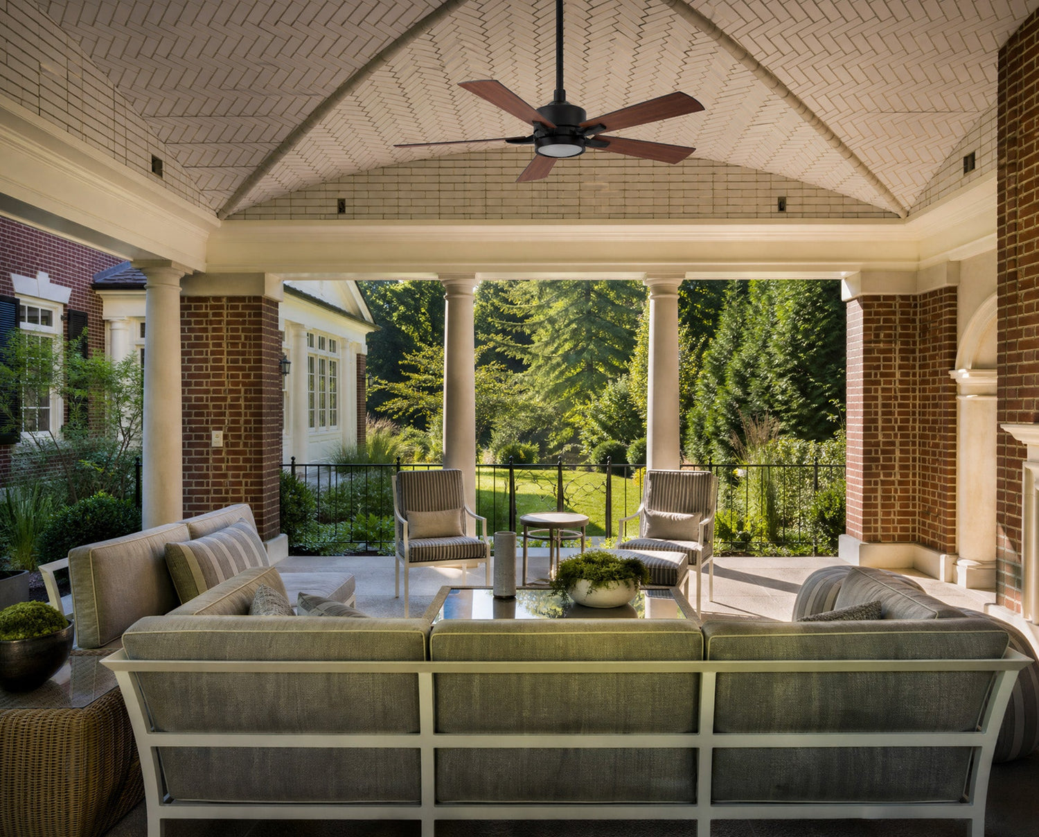 Matte black ceiling fan with light fixture and five medium-toned wood blades installed on a vaulted herringbone ceiling. The outdoor ceiling fan anchors a luxury covered patio space complete with comfortable outdoor furniture, brick pillars, and backyard views.