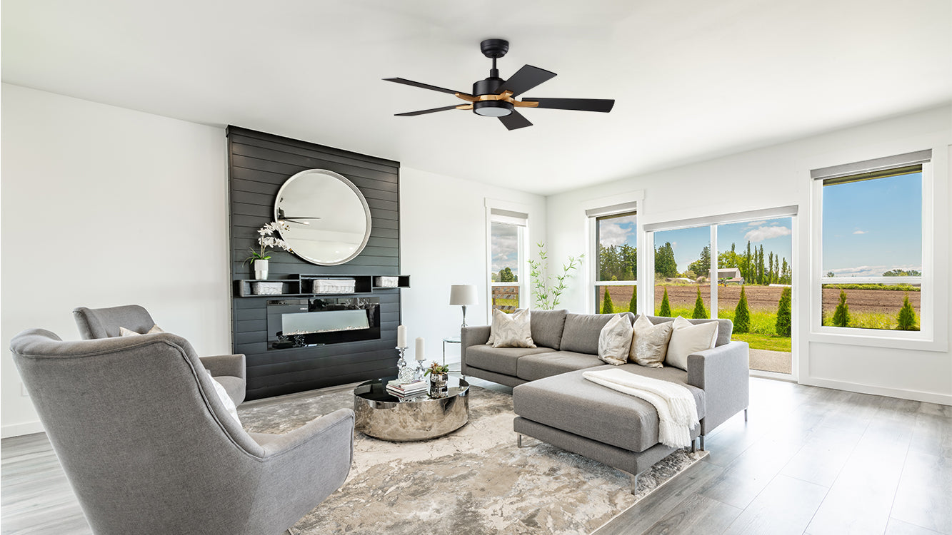 A modern living room featuring an Apex black and gold ceiling fan with a light fixture. The room has light gray walls and flooring, with large windows providing a view of a green landscape. The seating area includes a gray sectional sofa with various throw pillows, a matching armchair, and a round silver coffee table on a textured area rug. A black accent wall with a fireplace and a large round mirror above it adds a sophisticated touch.