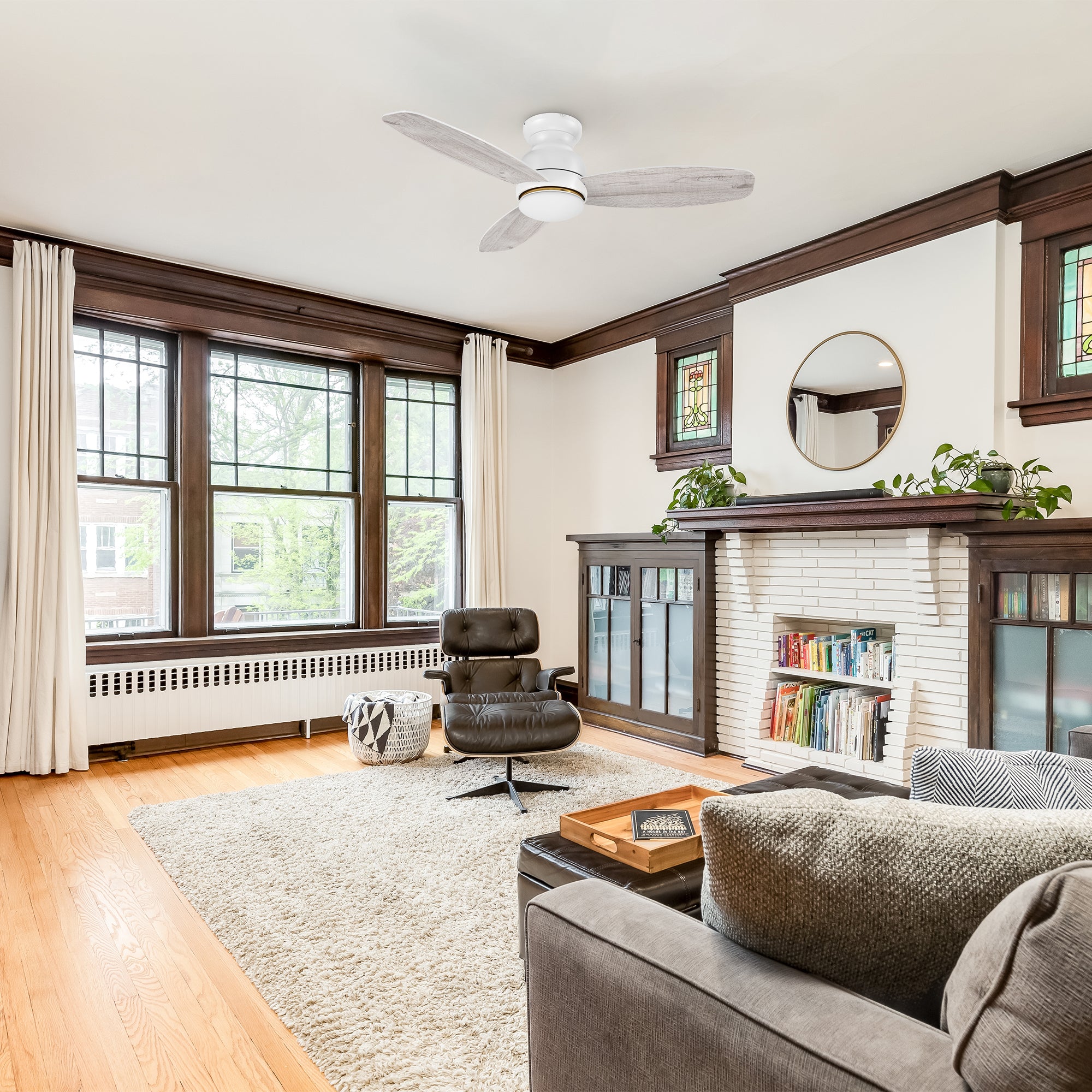 Modern living room featuring the Arran 48-inch flush mount ceiling fan with an integrated LED light kit. The fan includes three light wood-finished blades and a minimalist white housing, seamlessly blending with the bright and airy decor. The room showcases large windows, dark wood trim, a white brick fireplace with built-in cabinets, and comfortable seating for a cozy yet sophisticated ambiance. 