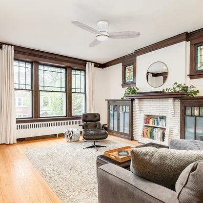 Modern living room featuring the Arran 48-inch flush mount ceiling fan with an integrated LED light kit. The fan includes three light wood-finished blades and a minimalist white housing, seamlessly blending with the bright and airy decor. The room showcases large windows, dark wood trim, a white brick fireplace with built-in cabinets, and comfortable seating for a cozy yet sophisticated ambiance. 