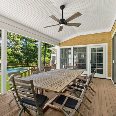 Aspen 48-inch outdoor smart ceiling fan with light in black finish, installed on a covered balcony with wood deck and patio dining set, overlooking a backyard with green trees and a swimming pool.