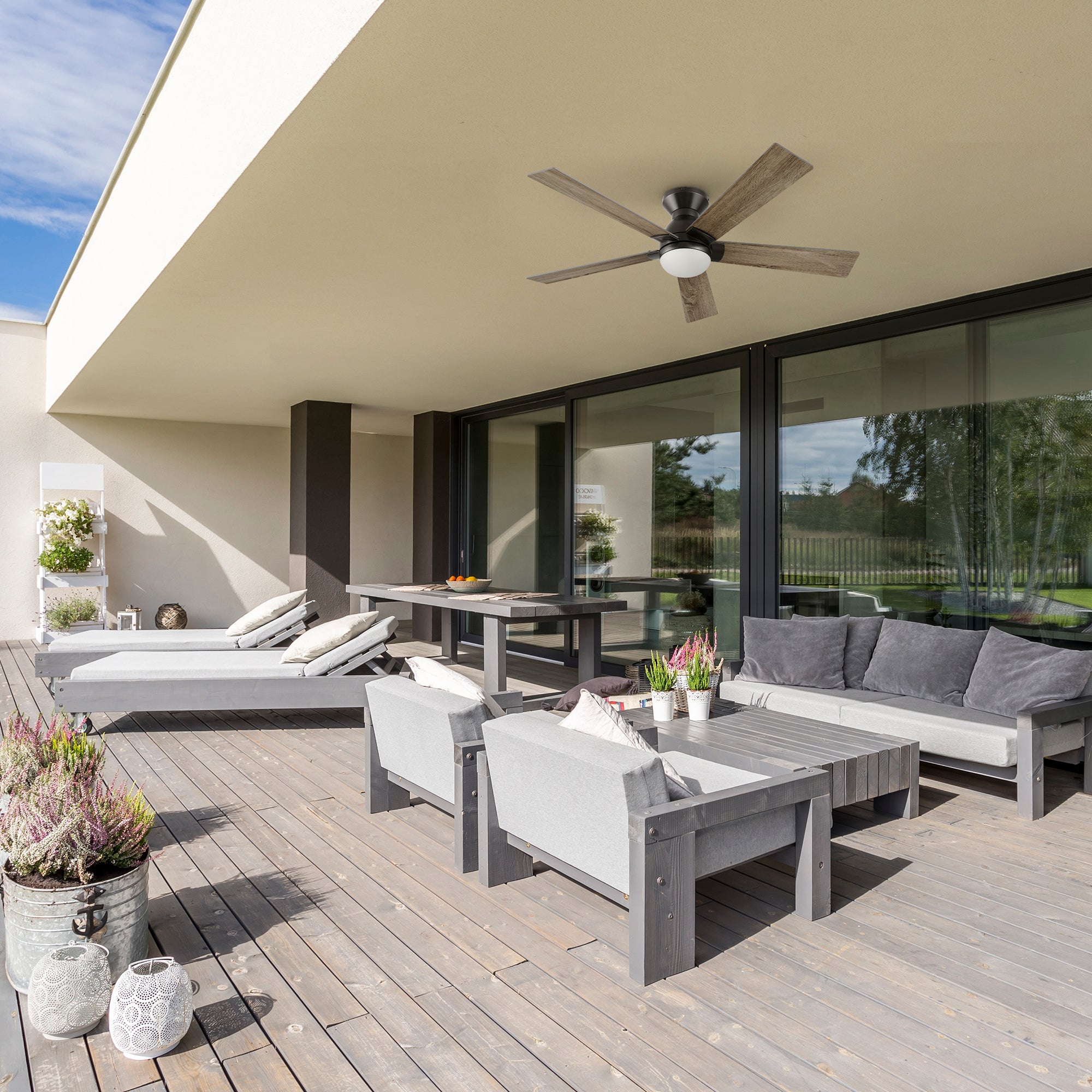 A stylish covered patio showcases the Aspen 52" Flush Mount Ceiling Fan with light and smart controls. With light wood blades and a sleek design, it pairs perfectly with the wood deck, cozy gray seating, and lush potted plants—delivering powerful airflow and modern comfort for outdoor living. 