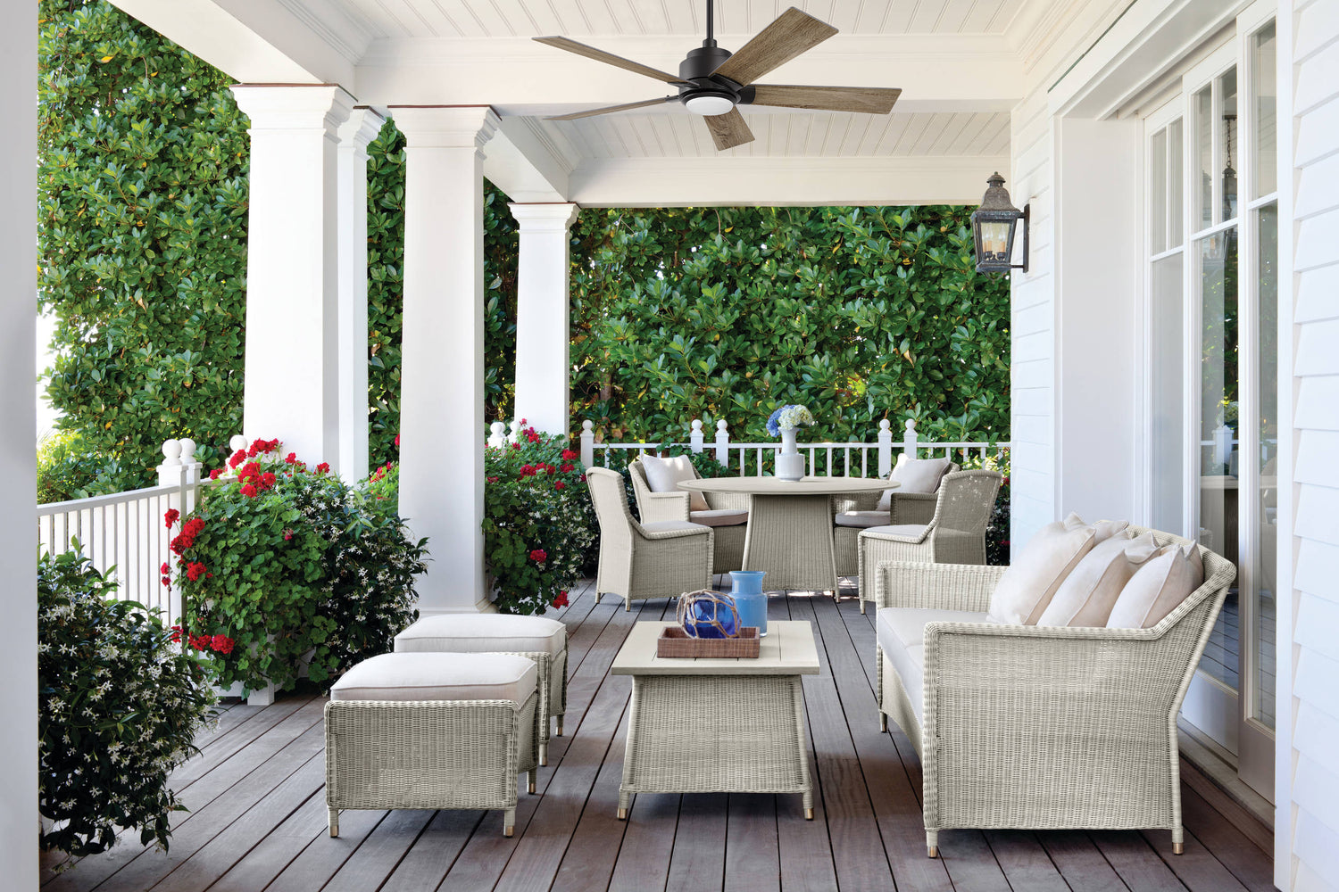 A Memorial Day 2025 banner featuring a covered outdoor patio with a white Aspen smart ceiling fan with LED light, mounted on a wooden ceiling. The fan has five blades and illuminates a cozy seating area with cushioned wicker chairs, a glass-top coffee table with a vase of flowers, and a beige rug, set against a backdrop of greenery. The banner includes red, white, and blue accents with text promoting a Memorial Day sale.