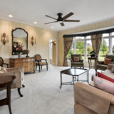 A traditional living room with light-colored, striped wallpaper features a modern, low-profile 52-inch dark wood ceiling fan with an integrated LED light as the focal point. The space is furnished with an antique-style wood dresser beneath a large ornate mirror, multiple upholstered accent chairs in shades of beige and green, and a dark metal octagonal coffee table. A large bay window with decorative patterned curtains overlooks a green lawn. #color_wood