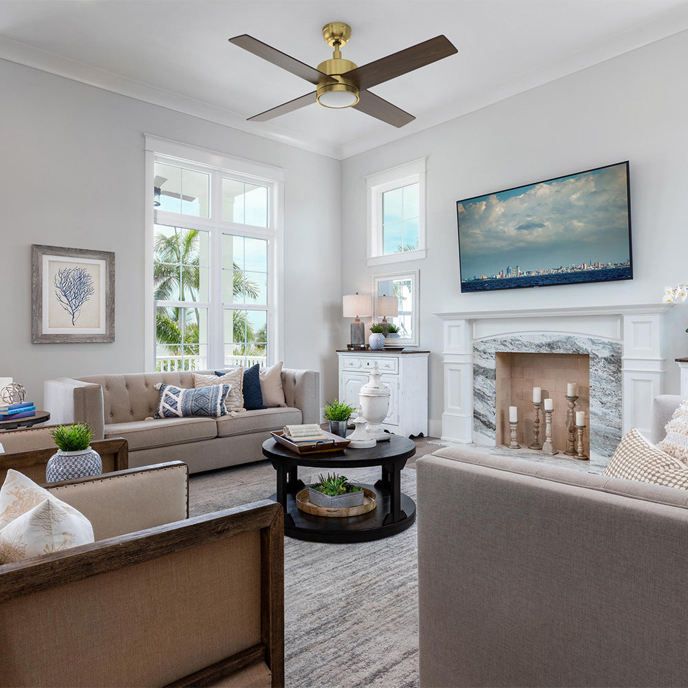 A spacious, sunlit living room is furnished with two beige sofas and two wood-framed armchairs arranged around a round black coffee table. Centered in the room is a 52 inch Smafan Rossendale gold ceiling fan with light, featuring four dark wood blades and a sleek gold motor hub. The room&