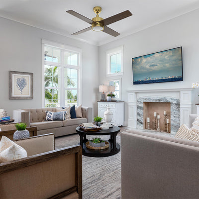 A spacious, sunlit living room is furnished with two beige sofas and two wood-framed armchairs arranged around a round black coffee table. Centered in the room is a 52 inch Smafan Rossendale gold ceiling fan with light, featuring four dark wood blades and a sleek gold motor hub. The room's focal point is a white fireplace with a gray marble surround, topped by a large flat-screen TV. Large windows and a glass-paneled door offer a view of palm trees and water, enhancing the coastal aesthetic. #color_gold