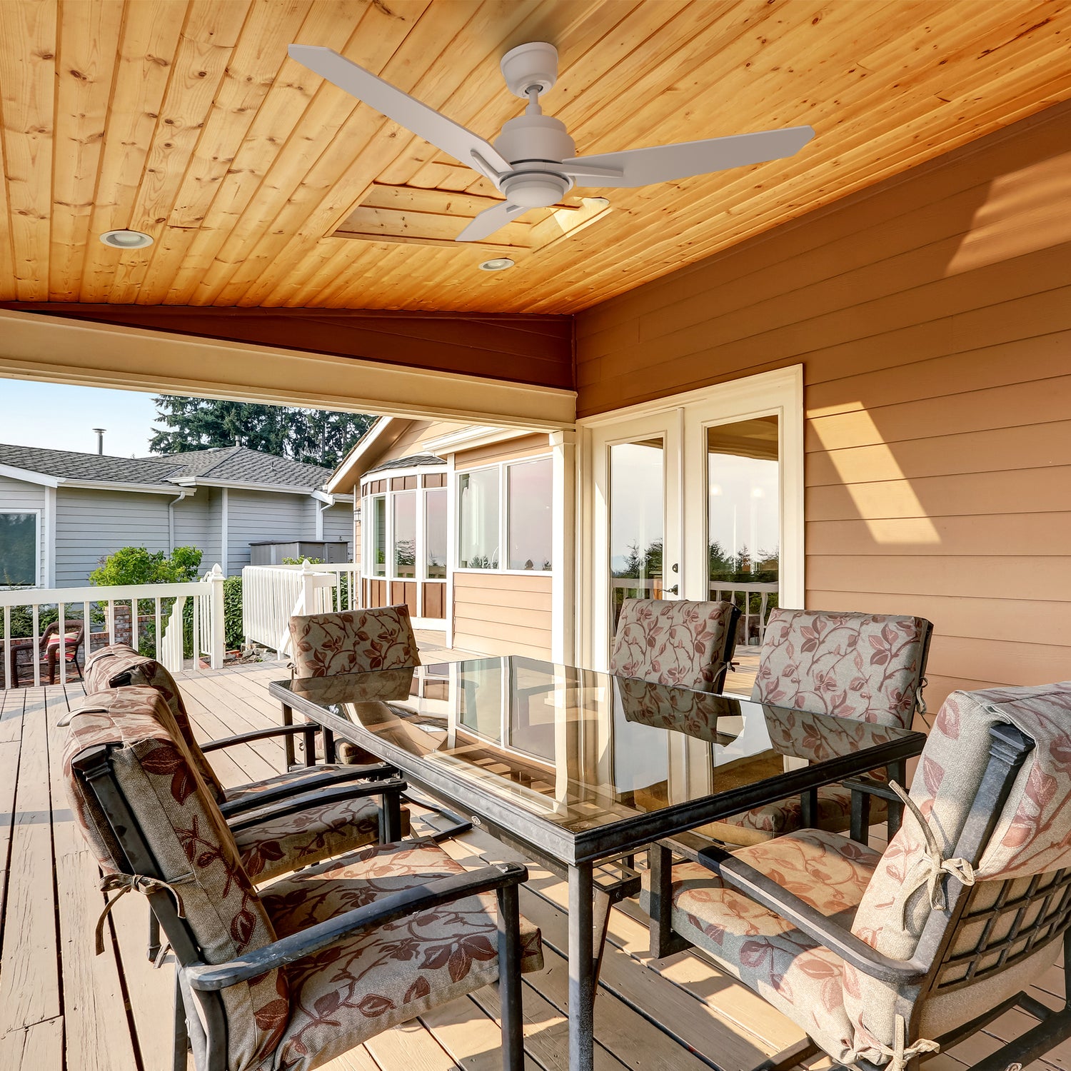 A modern covered patio featuring a 48-inch Carro Smafan Tilbury outdoor smart ceiling fan with LED light and remote, mounted on a white ceiling.   The fan, with three sleek blades in a white finish, illuminates a cozy seating area with cushioned wicker chairs, a glass-top coffee table.  The patio overlooks a scenic backyard with greenery and wooden fencing, showcasing the fan&
