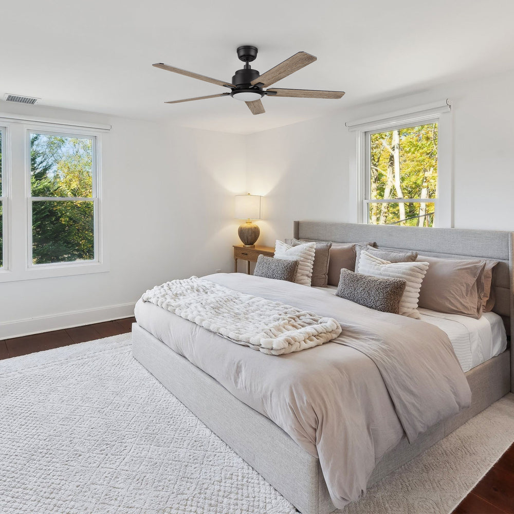 Carro Smafan Essex2 52-inch black smart ceiling fan with LED light installed in a cozy, modern bedroom with wood flooring, large windows, and neutral-colored bedding. 