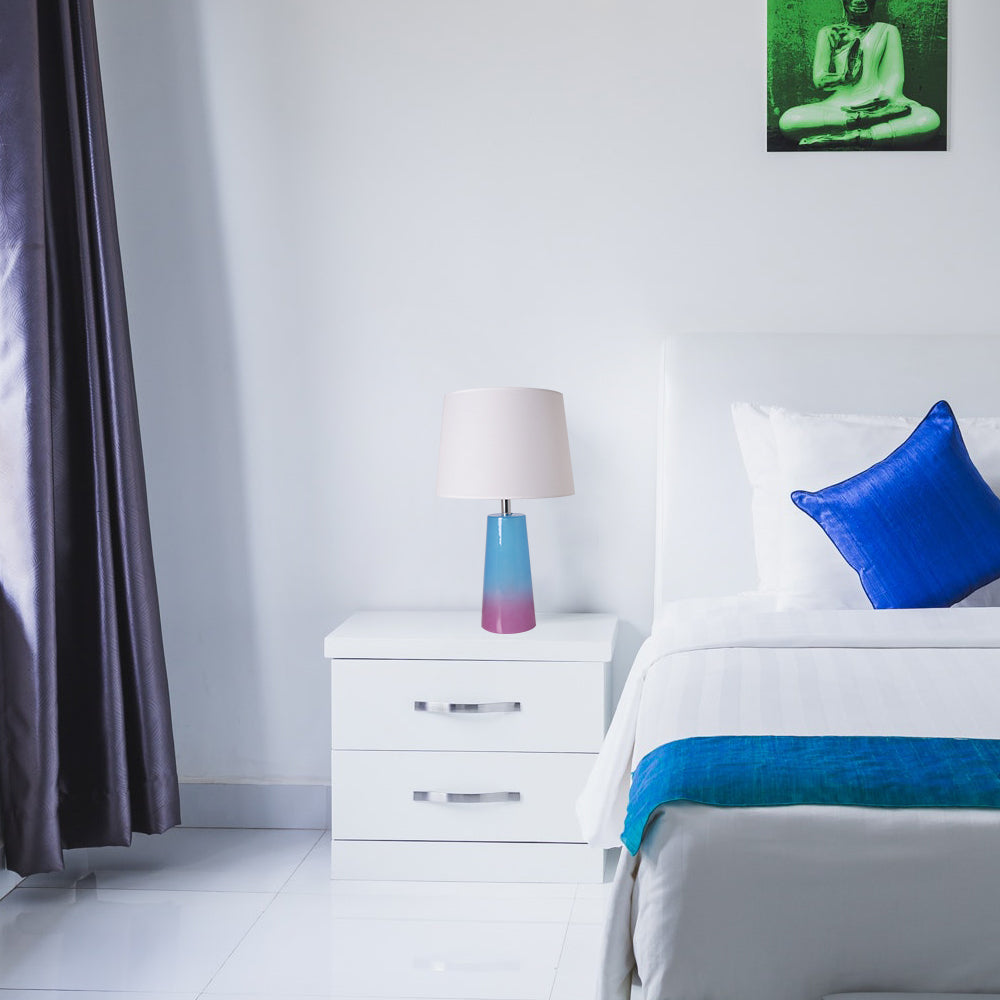 The table lamp placed on a white nightstand next to a bed with white bedding and blue accents in a minimalist, bright room with gray curtains. 