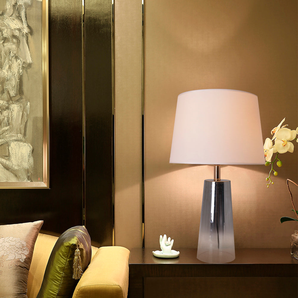 A side table view of a Carro Smafan Rosemary Table Lamp, 25 inches tall, with a Chrome Grey Ombre finish and white shade, placed on a table in a cozy corner. The lamp is shown illuminating the space with a soft glow, highlighting its stylish design and lighting functionality. 