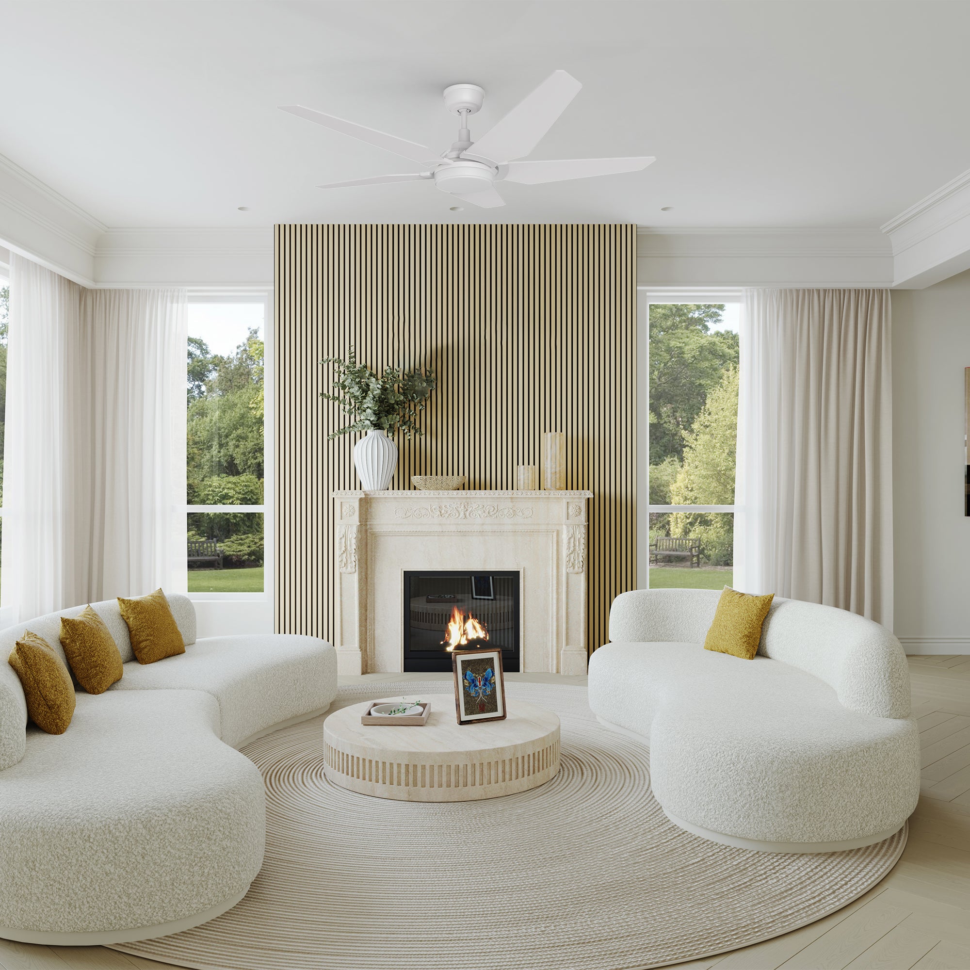 A sophisticated and airy living room showcases the Carro Smafan Voyager 52” outdoor smart ceiling fan in white with an integrated LED light and remote. The room features curved white boucle sofas with mustard accent pillows, a modern fireplace with a slatted wood panel backdrop, and large windows framing a lush green outdoor view. Neutral tones and soft textures complete the serene, upscale aesthetic. 