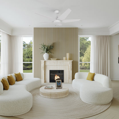 A sophisticated and airy living room showcases the Carro Smafan Voyager 52” outdoor smart ceiling fan in white with an integrated LED light and remote. The room features curved white boucle sofas with mustard accent pillows, a modern fireplace with a slatted wood panel backdrop, and large windows framing a lush green outdoor view. Neutral tones and soft textures complete the serene, upscale aesthetic. 