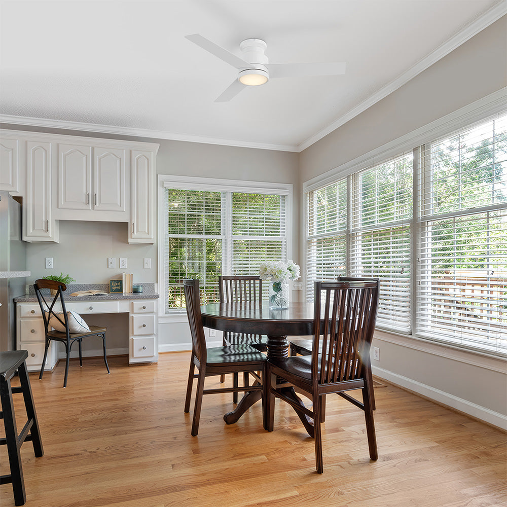 A bright modern dining area featuring the Carro Smafan Ariccia 52-inch low profile ceiling fan in white, with an integrated LED light that illuminates the space. Large windows, hardwood floors, and dark wood dining furniture create an airy, inviting setting. 