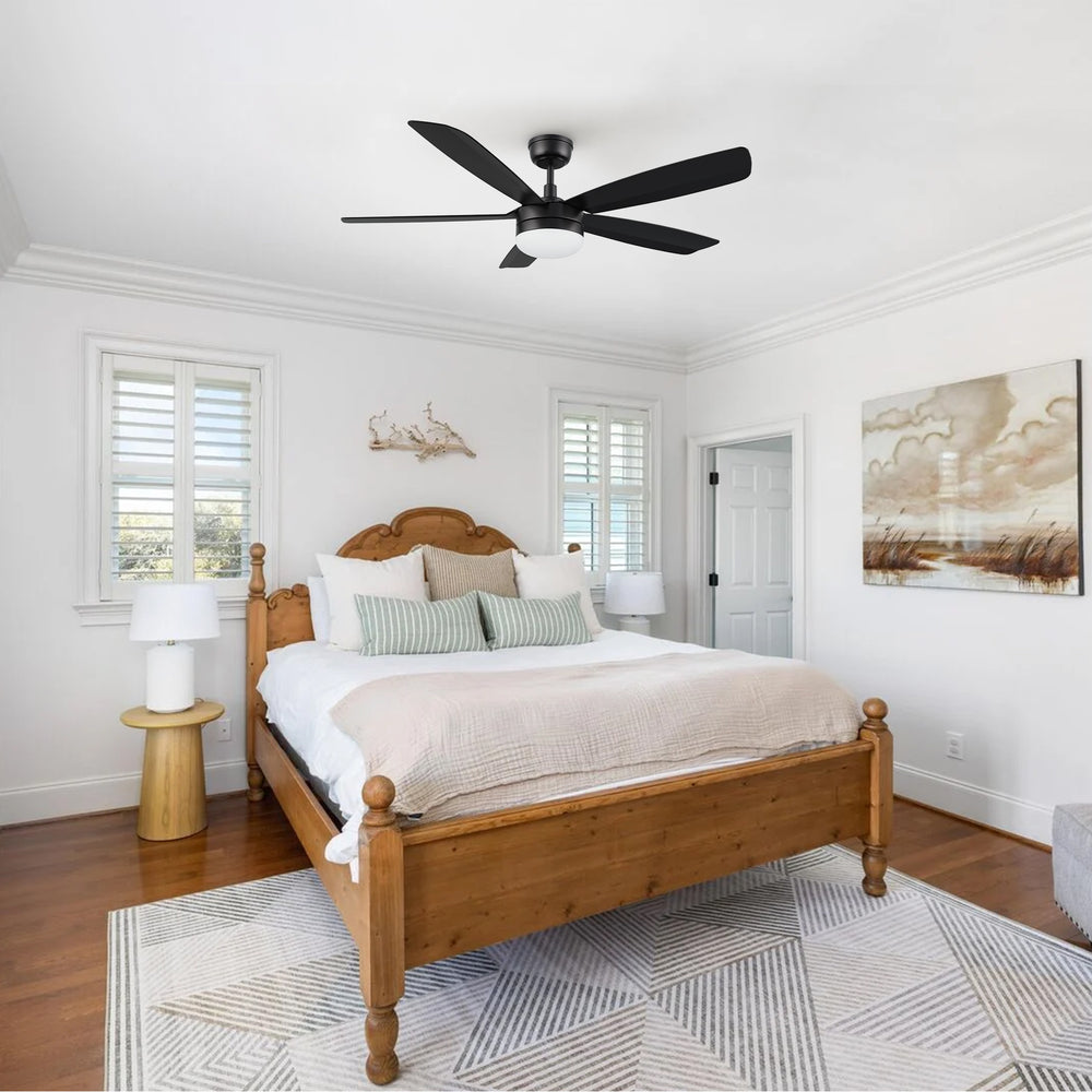 A stylish 52-inch black ceiling fan with a central light fixture installed in a modern bedroom with minimalist decor, featuring a low platform bed and large windows. 