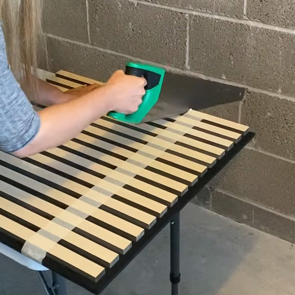 The same person cuts a wooden slat wall panel with a green-handled saw on a table, set against a concrete block wall.