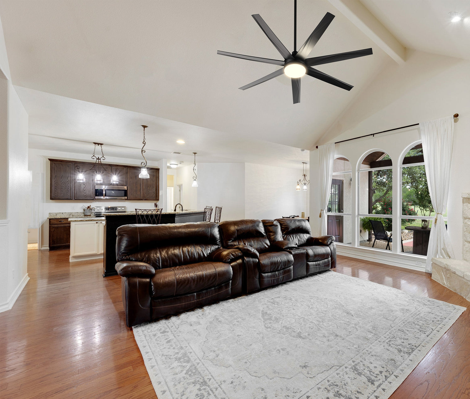 Open-concept living room and kitchen with hardwood floors, a vaulted ceiling, a large brown leather reclining sectional sofa, and a modern black seven-blade ceiling fan