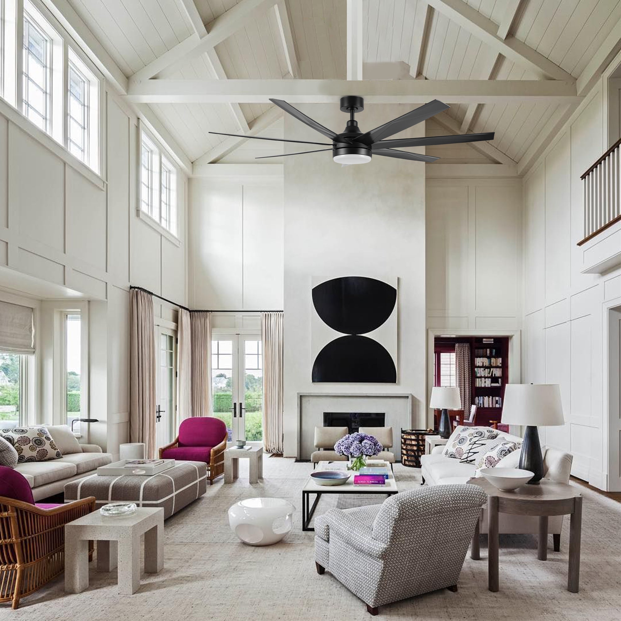 A large living room with high ceilings and white walls, a sleek black ceiling fan above the fireplace, contemporary furniture in neutral tones with pops of purple accents, and natural light pouring through the windows. 