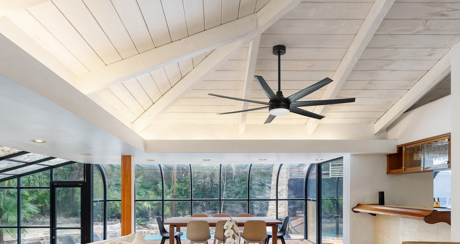 A wide, bright room with a high, vaulted white paneled ceiling from which a large, dark modern ceiling fan hangs.Daventry Best High CFM 65-inch matte black ceiling fan with 7 sleek blades and integrated dimmable LED light, designed for large indoor spaces, controlled by a remote, enhancing airflow and providing versatile lighting. The room features a dining area with a long table and chairs situated in front of expansive glass windows and doors that offer a view of lush greenery or a pool area outside.