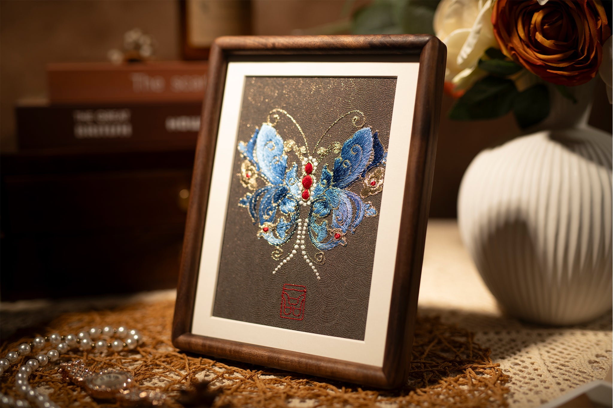 A wood-framed embroidered artwork showing beautiful blue butterfly, paired with flowers and jewelry and scented candles, is placed on the tabletop.