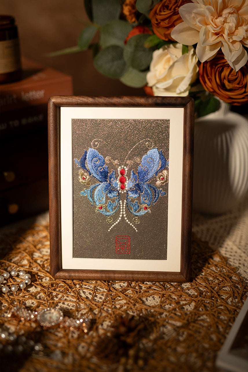 A wood-framed embroidered artwork showing beautiful blue butterfly, paired with flowers and jewelry and scented candles, is placed on the tabletop.
