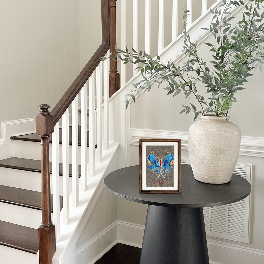 An embroidered tabletop artwork of butterflies and a potted willow plant are placed on a side table next to the stairs.