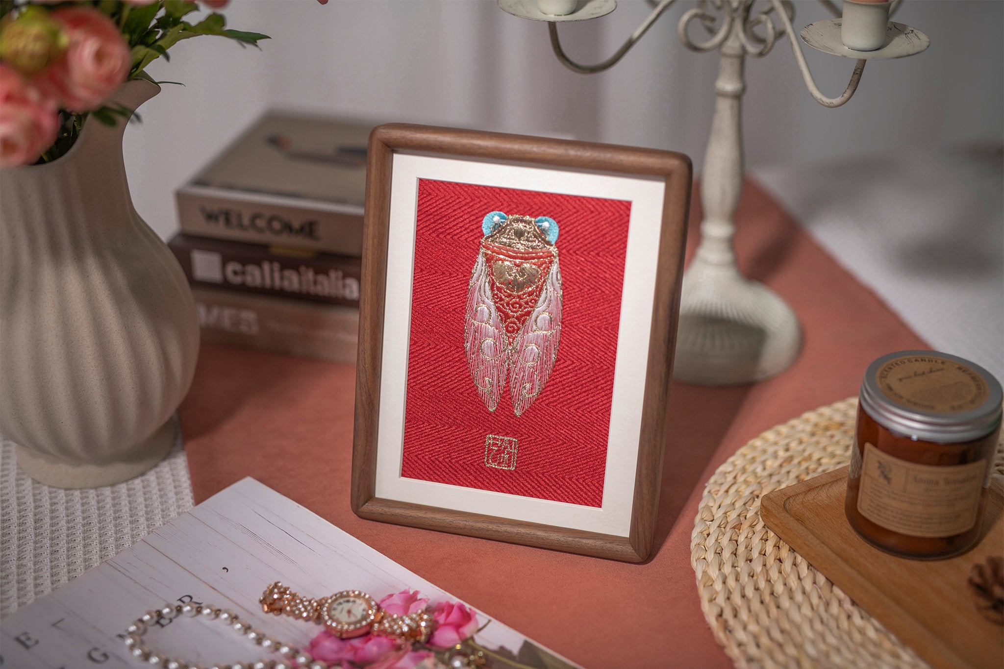 The golden Cicada embroidery artwork, paired with a five-head candlestick, pink flowers, jewelry, and scented candles, is quietly placed on top of a wood-grain table.