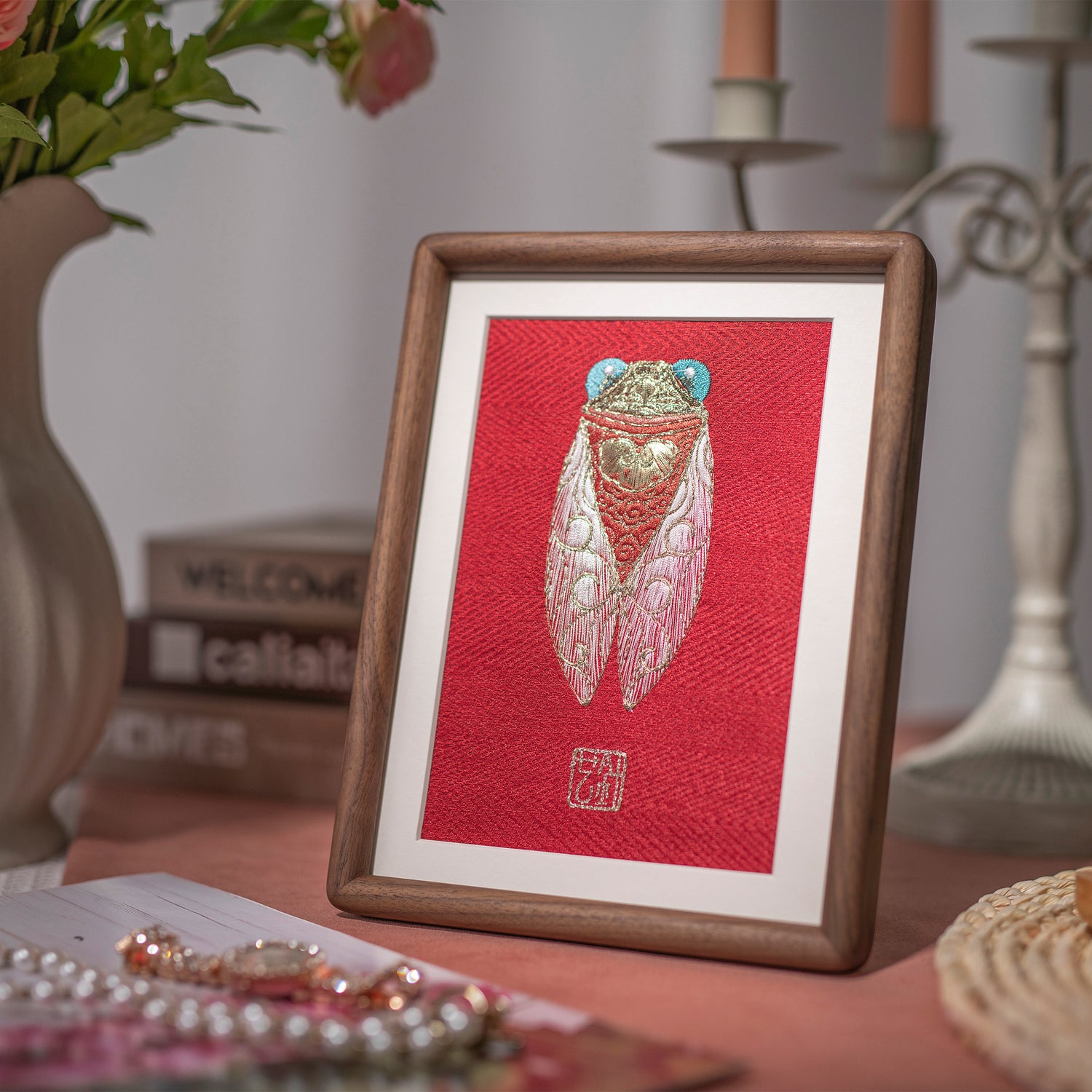 The golden Cicada embroidery artwork, paired with a five-head candlestick, pink flowers, and jewelry, is quietly placed on top of a wood-grain table