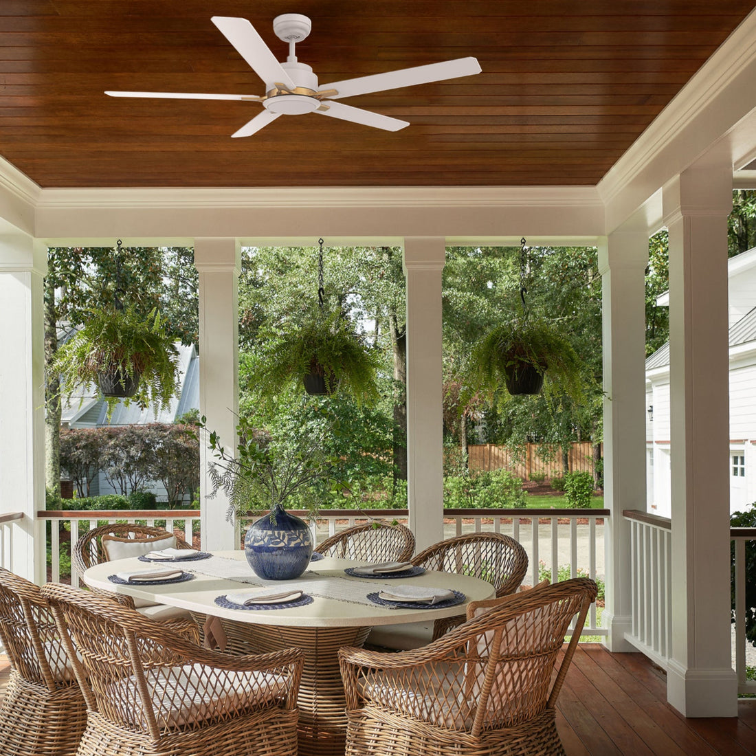 A covered patio with a wooden ceiling features the Essex 52" White Outdoor Ceiling Fan with gold-accented blades. Paired with a wicker dining set and lush greenery, it brings cool comfort and elegant style to outdoor living.
