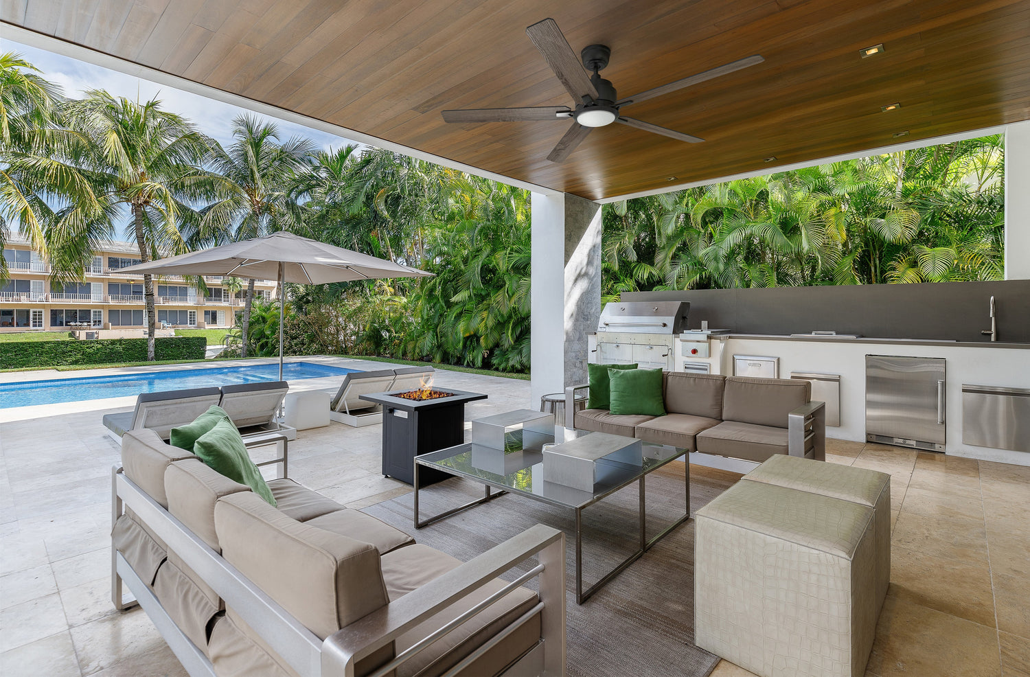 Modern covered outdoor kitchen and lounge area featuring the Essex 60-inch smart outdoor ceiling fan with light, mounted on a wood-paneled ceiling.  The space includes sleek patio furniture, a fire pit, built-in stainless steel appliances, and a view of a lush garden and swimming pool—perfect for Earth Day comfort and energy efficiency.