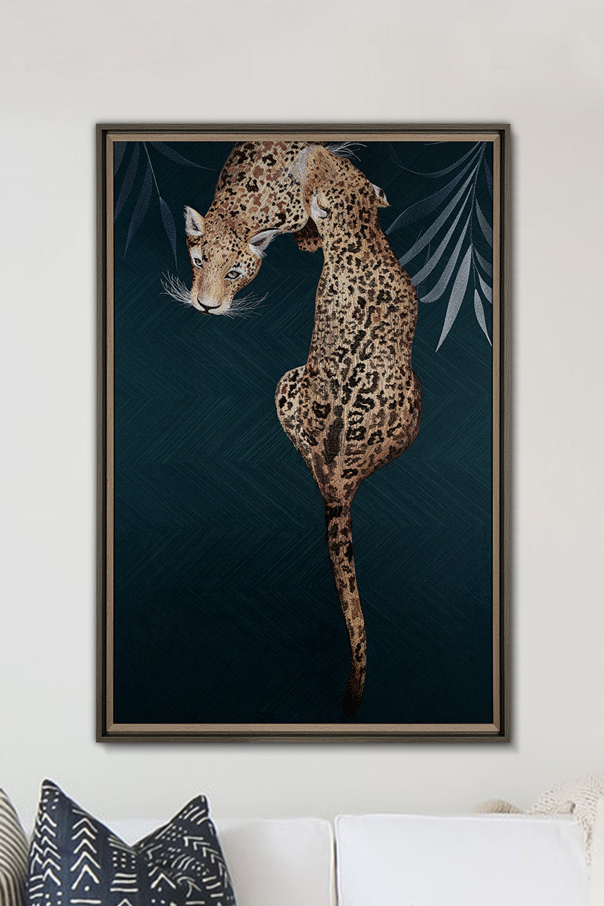 A piece of embroidered art with two leopards snuggling together, flanked by palm leaves, and dominated by a dark green background, hangs on the wall.