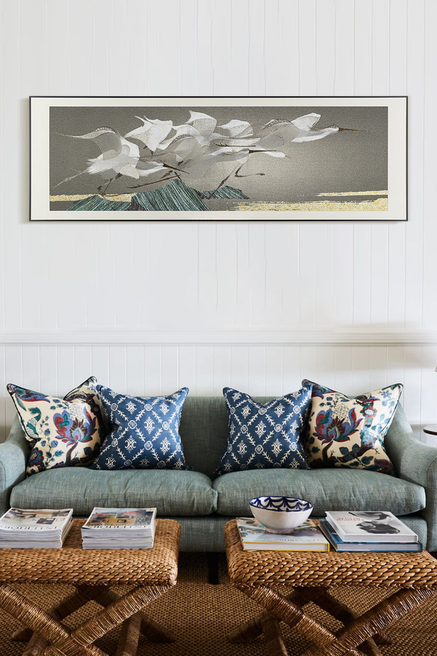 A framed embroidery wall art piece depicting egrets flying over mountains, displayed above an elegant sofa with blue and white patterned cushions in front of it.    The room features wicker coffee tables adorned with magazines and vases, all set against pristine white panelled walls.
