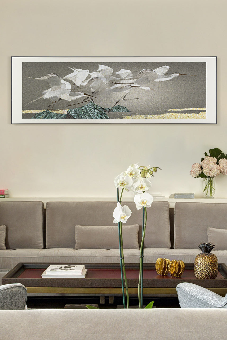 A large white decorative embroidery in the Chinese style, with a long frame depicting flying egrets. And three orchids in the foreground on a table top, with a light gray sofa in the background.