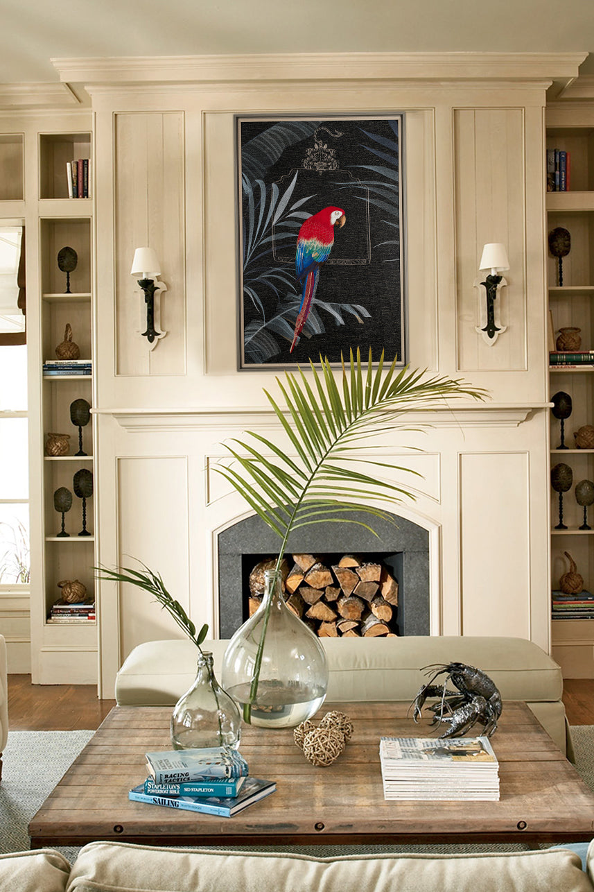 A coastal living room with light cream colored walls and white trim. In the center of the background is an embroidery wall art of a parrot sitting on a palm leaf in a black frame above the stone fireplace. On the coffee table are books and a large glass vase. A small metal sculpture and a wooden shelf holding books are also present. Wooden side tables add blue accents to the space, giving it a beach vibe. Natural lighting brightens the room.