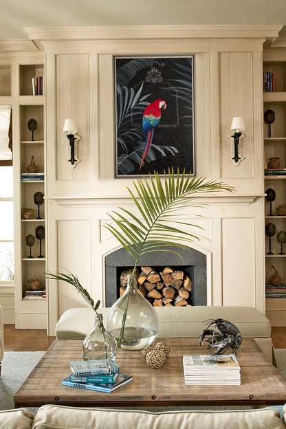 A coastal living room with light cream colored walls and white trim. In the center of the background is an embroidery wall art of a parrot sitting on a palm leaf in a black frame above the stone fireplace. On the coffee table are books and a large glass vase. A small metal sculpture and a wooden shelf holding books are also present. Wooden side tables add blue accents to the space, giving it a beach vibe. Natural lighting brightens the room.