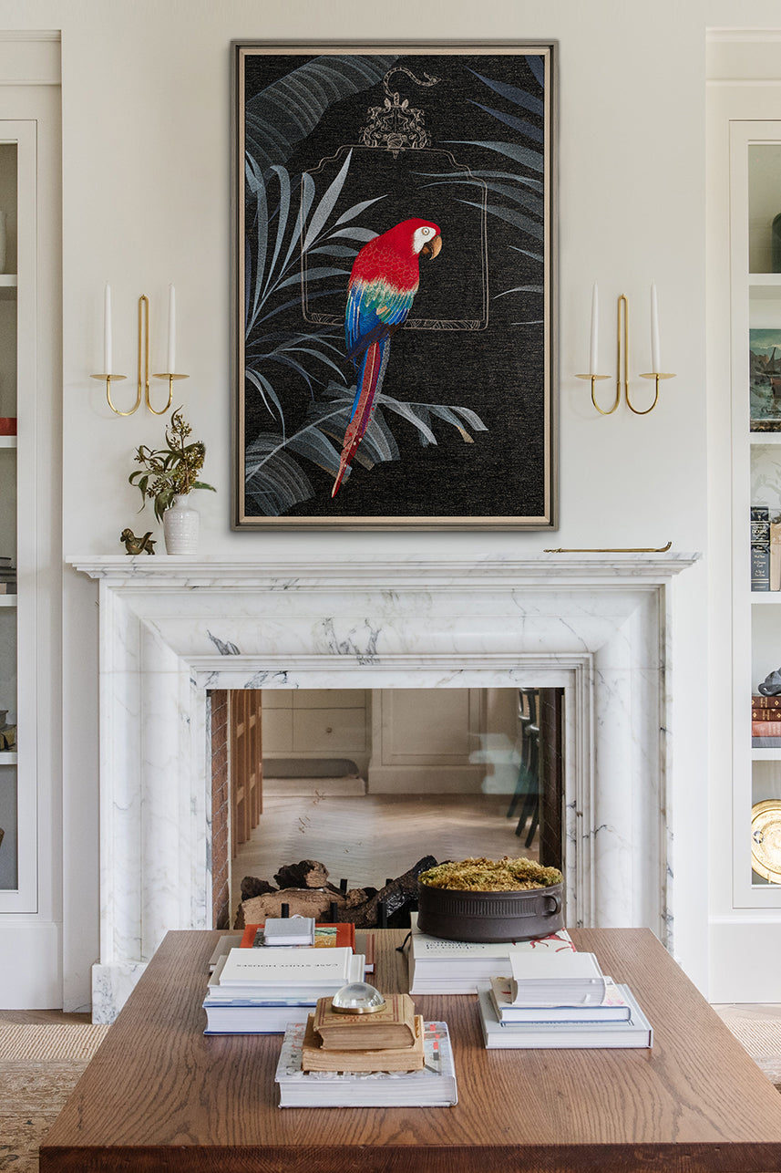 A photo of large framed embroidery art with red and blue macaw on it, above the fireplace in an elegant living room, coffee table books, marble shelf, brass sconces, a stack of luxury fashion magazines.