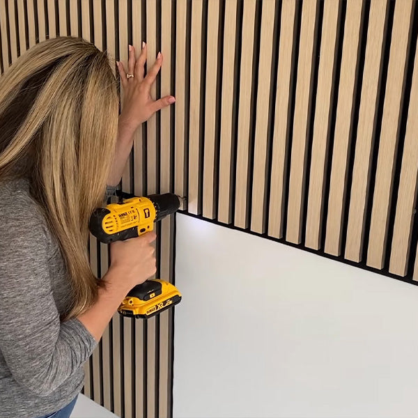 The person drills a wooden slat wall panel into a white wall using a yellow power drill, securing the slats in place.