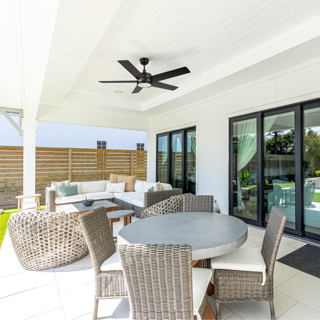 Lakeland 52-inch outdoor smart ceiling fan with LED light and remote, mounted on a white paneled ceiling above a stylish covered patio with wicker seating and a round dining table. 