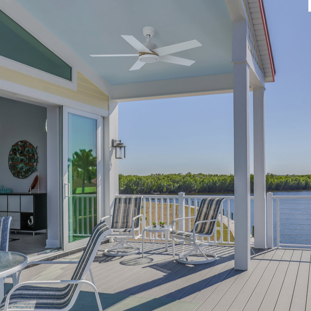 Bright waterfront patio featuring a white 52-inch Lakeland outdoor smart ceiling fan with integrated LED light, mounted on a light blue ceiling. The space includes striped lounge chairs and a table set, overlooking a scenic river and lush green shoreline. 