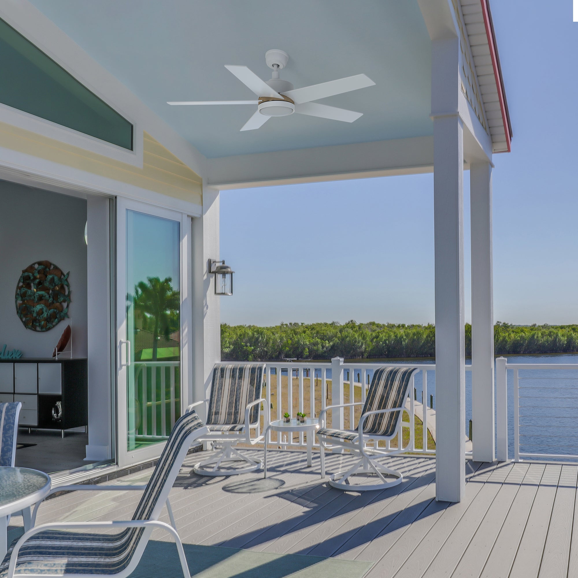 Bright waterfront patio featuring a white 52-inch Lakeland outdoor smart ceiling fan with integrated LED light, mounted on a light blue ceiling. The space includes striped lounge chairs and a table set, overlooking a scenic river and lush green shoreline. 