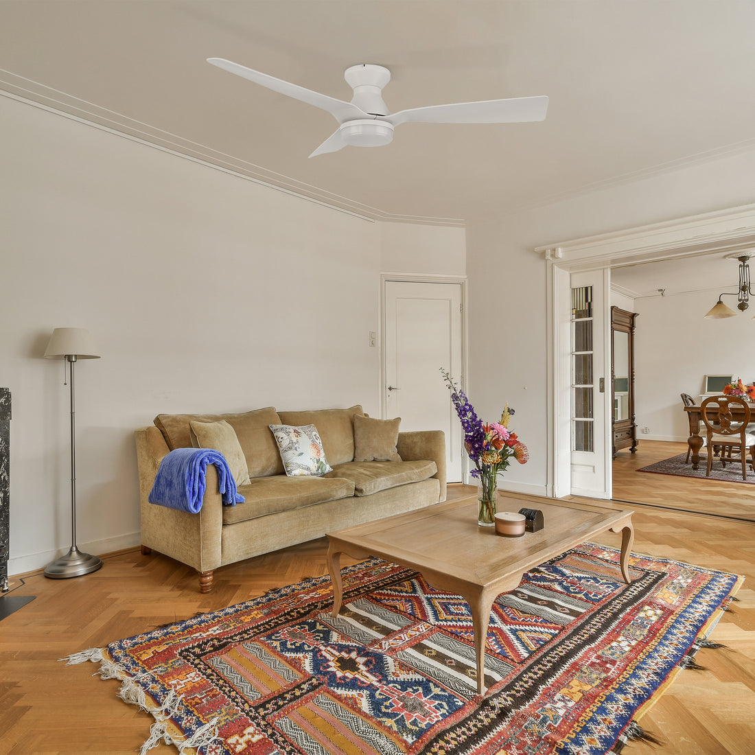 A cozy living room with a white flush mount ceiling fan featuring three curved blades and an integrated light. The space includes a beige sofa with a blue throw blanket, a wooden coffee table with a flower vase, and a colorful patterned rug on a herringbone wood floor. The room has an open layout leading to a dining area with vintage-style furniture. 