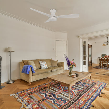 A cozy living room with a white flush mount ceiling fan featuring three curved blades and an integrated light. The space includes a beige sofa with a blue throw blanket, a wooden coffee table with a flower vase, and a colorful patterned rug on a herringbone wood floor. The room has an open layout leading to a dining area with vintage-style furniture. 