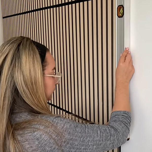 A person with long blonde hair, wearing a gray sweater, uses a level to measure and align a wooden slat wall panel on a white wall.