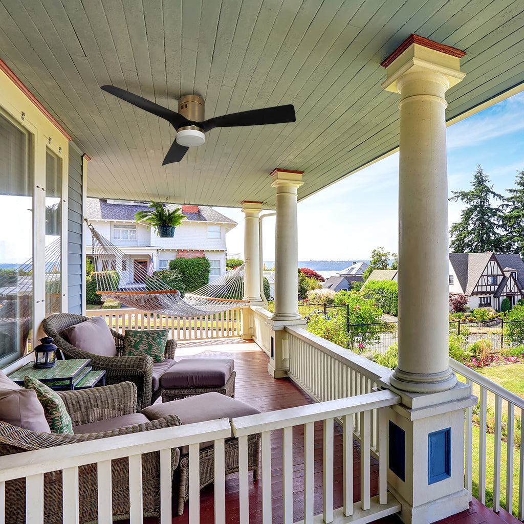 A brushed gold low-profile ceiling fan with three black blades is mounted on the light blue beadboard ceiling of a traditional covered porch. The spacious porch features large white columns, a matching railing, and two wicker lounge chairs with plush cushions and ottomans. In the background, a white hammock is strung across the deck, overlooking a scenic view of a coastal neighborhood and a large body of water under a bright sky. 