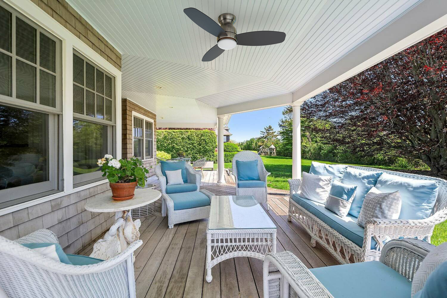A cozy covered patio featuring white wicker furniture with light blue cushions, surrounded by lush greenery and trees. At the center of the ceiling is a sleek, modern flush-mount ceiling fan (Trendsetter model) in a brushed nickel finish with dark blades. The setting evokes a serene and breezy outdoor retreat, perfect for a Mother’s Day moment of relaxation.