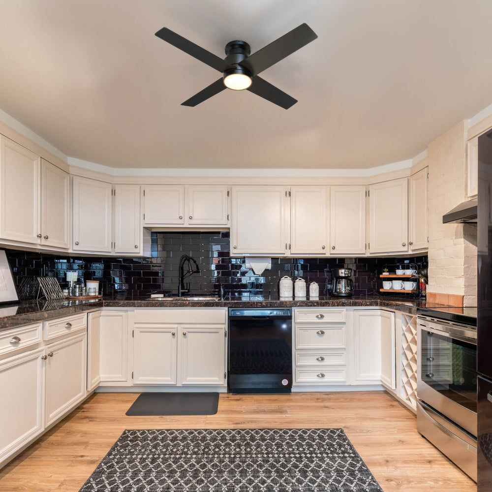 A modern kitchen with white cabinets, dark tile backsplash, and 52 inch black flush-mount ceiling fan with an integrated LED light in the center of the ceiling. The kitchen features stainless steel appliances, dark countertops, and a patterned area rug on a wooden floor. 