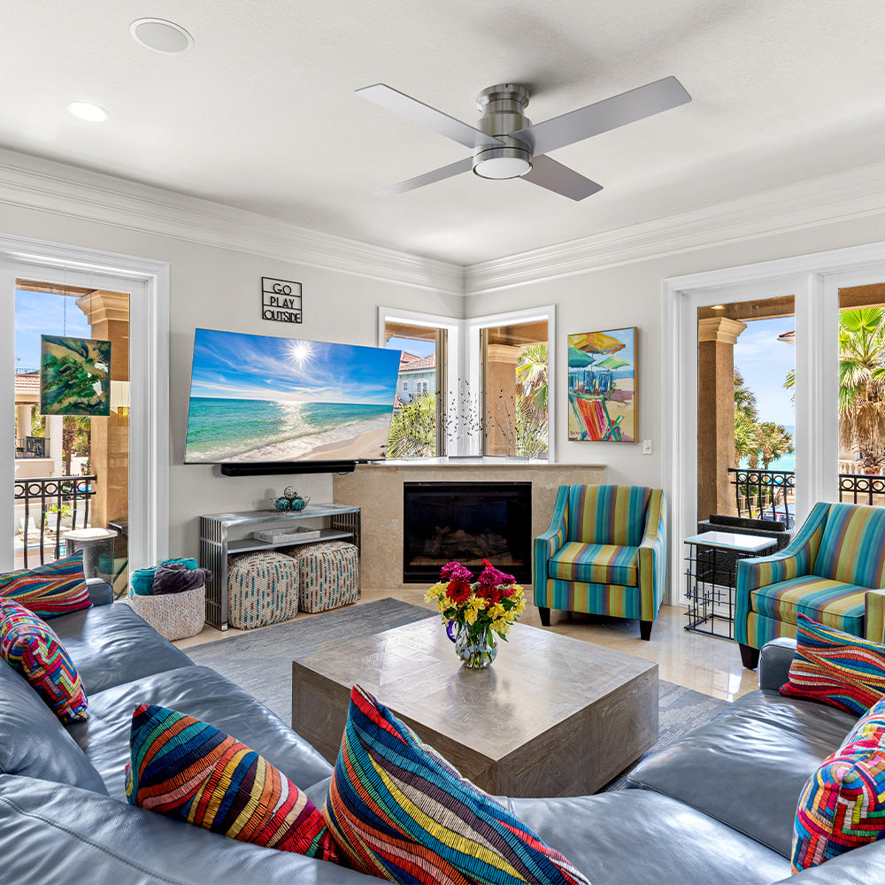 Coastal-inspired living room with a large gray sectional sofa, colorful patterned accent pillows, and striped armchairs facing a wall-mounted TV and flush mount ceiling fan with light. 