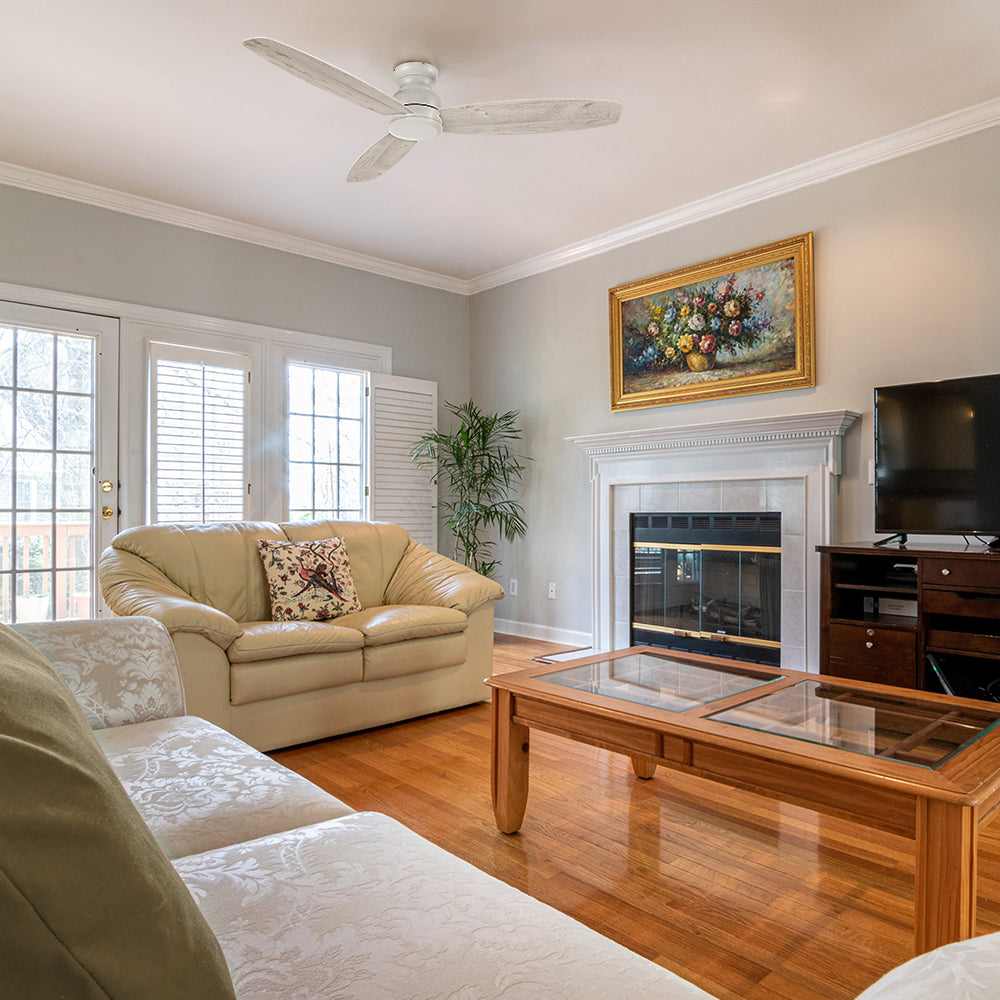 A cozy living room with a modern flush mount ceiling fan in white mounted on a beige ceiling. The space features a cream-colored loveseat, a matching floral-patterned sofa, and a glass-top wooden coffee table. A fireplace with a white mantel and a framed floral painting adds warmth to the room. A TV sits on a wooden entertainment center, and natural light streams in through large French doors with white shutters, complementing the light hardwood flooring. 