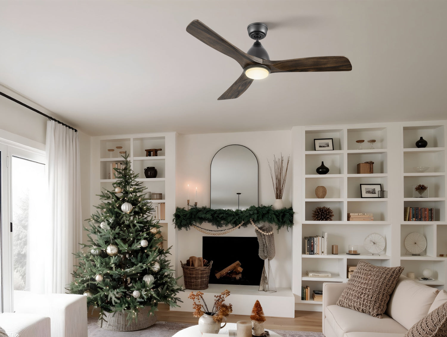 Cozy, modern living room decorated for Christmas, featuring a large Christmas tree with white lights and ornaments, a fireplace with a green garland, extensive white built-in bookshelves, neutral-toned furniture, and a dark wood three-blade ceiling fan.